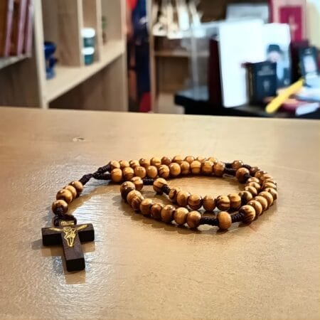 Χειροποίητο κομποσκόινι με ξύλινες χάντρες στο χρώμα της ελιάς και ξύλινο σταυρό με χρυσό Εσταυρωμένο, τοποθετημένο σε ξύλινη επιφάνεια